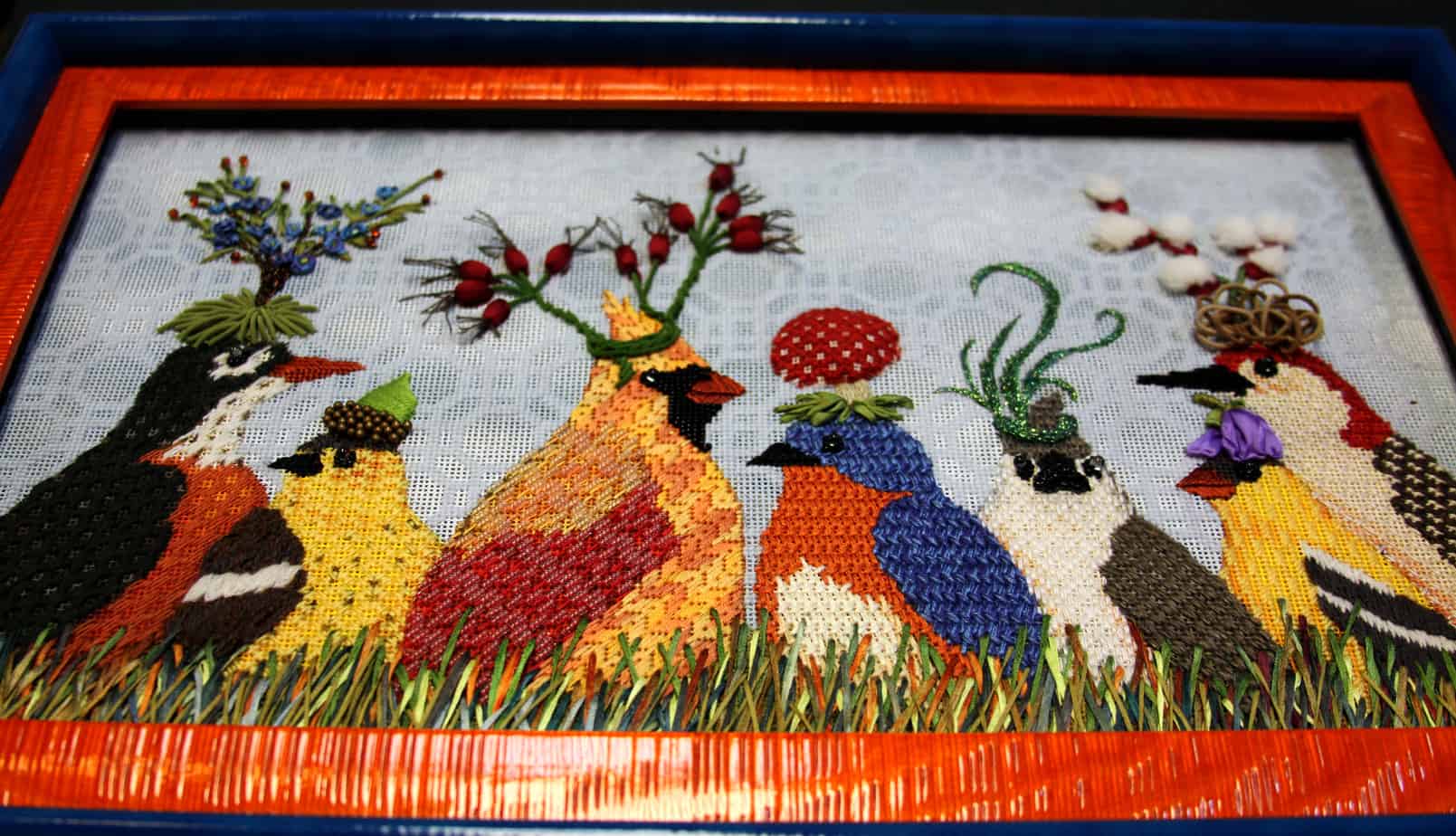 Needlepoint artwork of multicolored birds wearing plant-inspired hats.
