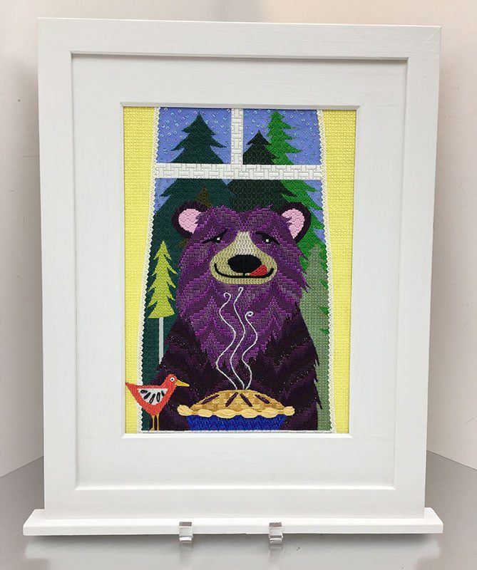 Framed needlepoint artwork of a purple bear smelling a pie with a bird beside said pie.